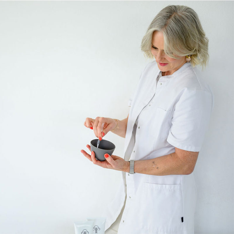 Ältere Frau in weißem Kittel bereitet Peptid Lifting Maske vor, hält Schale und Spatel in heller Umgebung​​​​​​​​​​​​​​​​​​​​​​​​​​​​​​​​​​​​​​​​