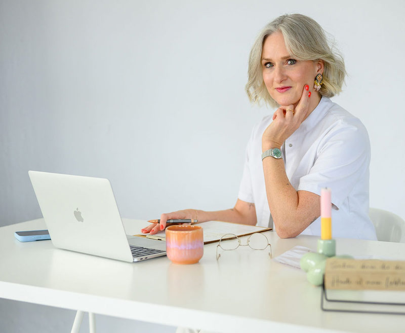 Ältere Frau in weißem Kittel sitzt am Schreibtisch mit Laptop, Notizblock und Kerze in heller, ruhiger Umgebung​​​​​​​​​​​​​​​​​​​​​​​​​​​​​​​​​​