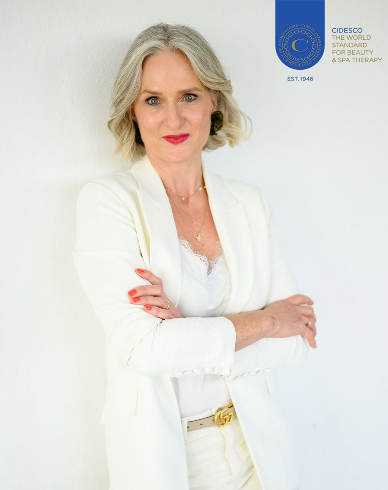 Porträt einer Frau mit blondem Haar im weißen Anzug vor weißem Hintergrund und CIDESCO Logo für Beauty und Spa Therapie.​​​​​​​​​​​​​​​​​​​​​​​​​