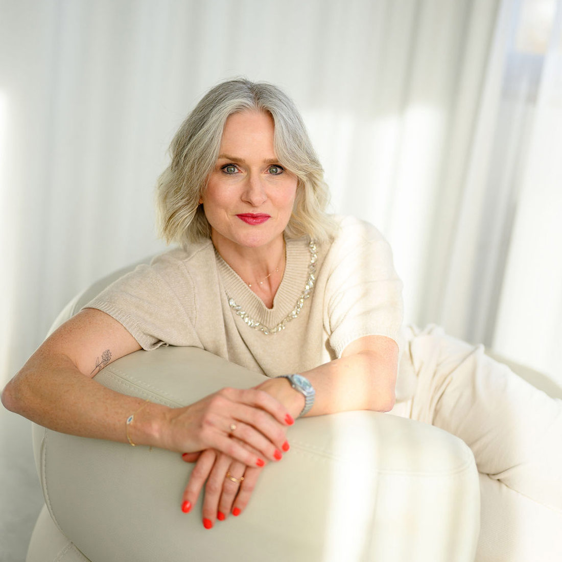 Ältere Frau mit grauen Haaren und rotem Nagellack sitzt entspannt auf weißem Sofa in heller, freundlicher Umgebung​​​​​​​​​​​​​​​​​​​​​​​​​​​​​​​